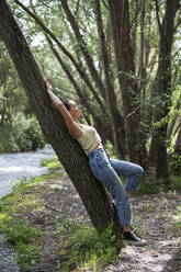 Woman leaning on tree trunk in forest - LMCF00304