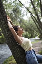 Woman with arms raised leaning on tree trunk in forest - LMCF00305