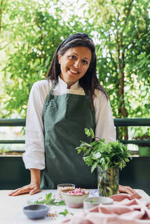 Smiling chef wearing apron standing near table in balcony - MEUF09153