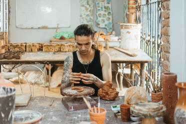 Focused young tattooed male artisan in black tank top working on clay wheel while shaping ceramic pot in professional workshop - ADSF44647