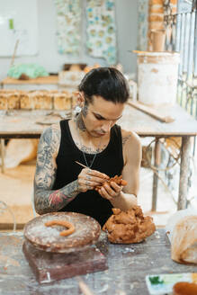 Concentrated young tattooed male potter in black tank top making clay product while working in professional workshop and creating earthenware - ADSF44648