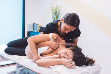 Young female osteopath using fingers and pressing shoulder part of pregnant patient lying on table during physiotherapy procedure in clinic - ADSF44652