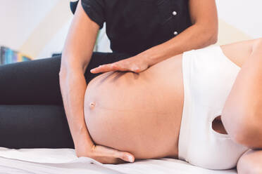 Crop osteopath treating pregnant patient and lying on table while treating baby in belly for correct position during session - ADSF44653