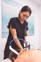 Young female therapist in uniform looking down and working on back of unrecognizable patient lying on belly on table with equipment attached in clinic - ADSF44660