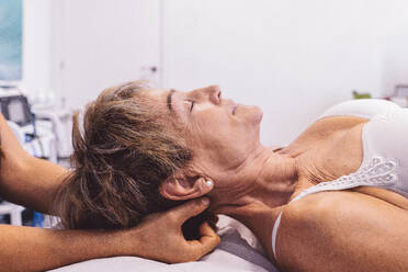 Side view of unrecognizable female therapist applying slow movements with fingers to sooth neck pain of senior lady lying on bed with eyes closed in clinic - ADSF44662