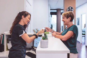 Side view of middle aged female patient standing at reception counter and laughing with lady receptionist while settling medical bills with credit card and looking at each other - ADSF44664