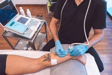 Unrecognizable doctor in latex gloves treating patient with ultrasound equipment while examining leg during appointment in contemporary hospital - ADSF44667