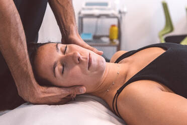 Unrecognizable professional osteopath massaging neck of female patient lying on table with closed eyes during appointment in modern clinic - ADSF44672