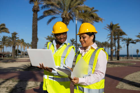 Builders in hardhat and engineer vest standing near construction site with laptop while discussing project - ADSF44702