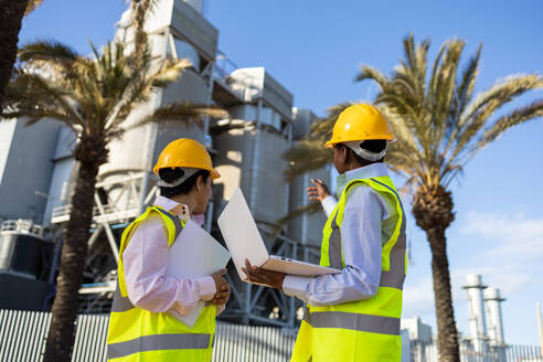 Low angle back view of unrecognizable builders in hardhat and engineer vest standing near construction site with laptop while discussing project - ADSF44703