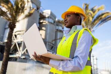 Low angle of young ethnic African America woman builder in uniform and helmet standing and using laptop while looking at project during work - ADSF44704