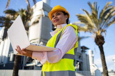 Low angle of young ethnic African America woman builder in uniform and helmet standing and using laptop while looking at project during work - ADSF44705