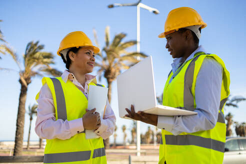 Low angle of builders in hardhat and engineer vest standing near construction site with laptop while discussing project - ADSF44708