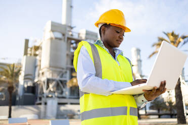 Low angle of young ethnic African America woman builder in uniform and helmet standing and using laptop while looking at project during work - ADSF44709