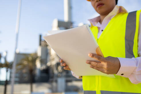 Young cropped unrecognizable woman in hardhat and engineer vest reading documents while standing on construction site during work process - ADSF44710