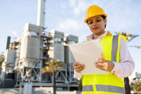 Young ethnic woman in hardhat and engineer vest reading documents while standing on construction site during work process - ADSF44711