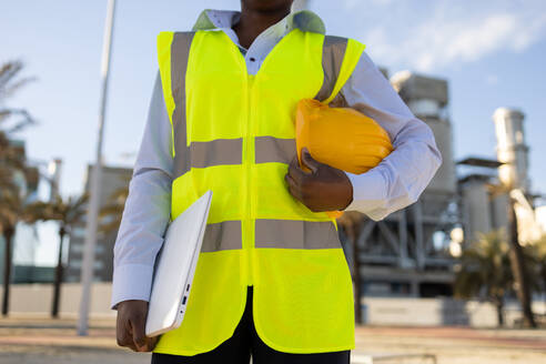 Crop anonymous African American female engineer in uniform and helmet and laptop standing on construction site during work - ADSF44712