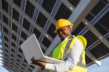 Low angle of happy African American female engineer in yellow vest and helmet standing with laptop while working on construction project with solar batteries looking at screen - ADSF44714