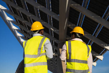 Back view of anonymous engineers in hardhats and uniform standing near installed solar panels on construction site - ADSF44715