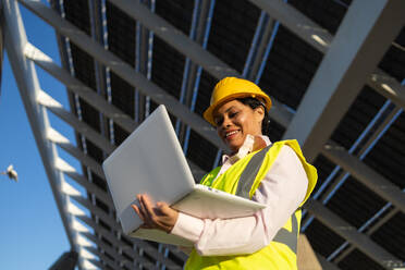 Low angle of happy African American female engineer in yellow vest and helmet standing with laptop while working on construction project with solar batteries looking at screen - ADSF44716