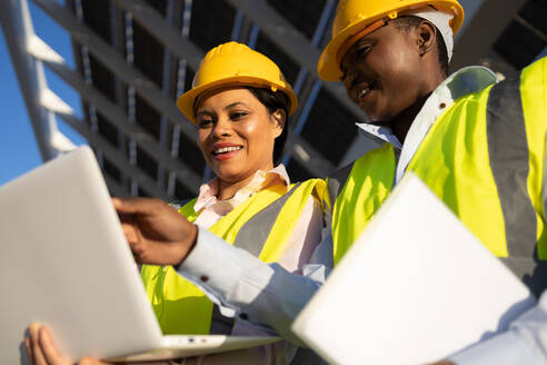 Low angle of young happy African American female builders in hardhat and uniform using laptop while working together and discussing project on construction site - ADSF44717