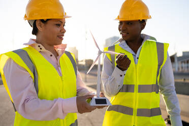African American female builders in hardhat and uniform using wind turbine while working together on construction site looking at each other - ADSF44718