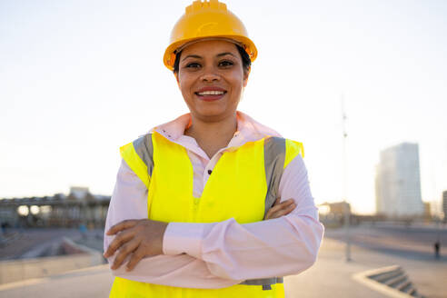 Positive young ethnic female engineer in hardhat and waistcoat smiling and looking at camera while standing on city street against cloudless sky - ADSF44720