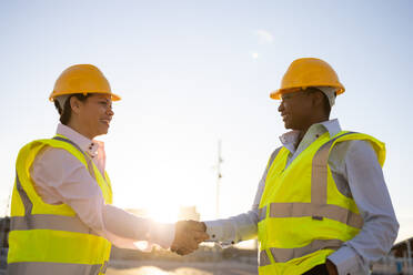 Side view of African American engineers in yellow waistcoat uniform happily shaking hands while standing looking at each other on city street over blurred landscape with sunlight - ADSF44721