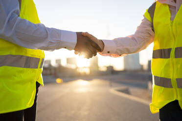 Faceless anonymous engineers in yellow waistcoat uniform shaking hands while standing on city street over blurred landscape with sunlight - ADSF44722