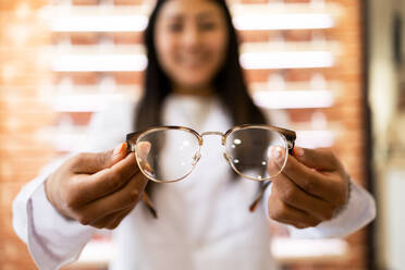 Blurred young eye doctor with long dark hair smiling and demonstrating optical glasses during work process - ADSF44723
