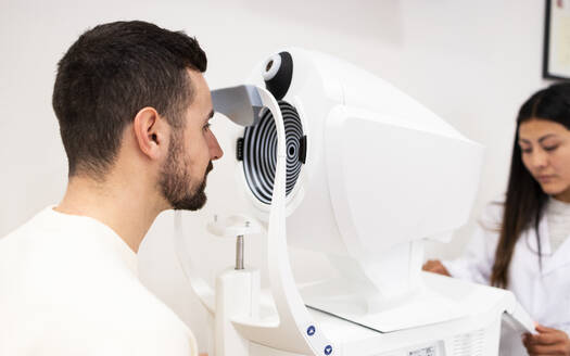 Side view of bearded male patient looking through optical phoropter during eye examining in medical clinic - ADSF44724