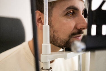 Side view of positive bearded male patient looking at slit lamp while visiting to ophthalmologist for examining vision in medical room - ADSF44725