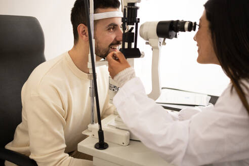 Side view of positive bearded male patient looking at slit lamp while visiting to ophthalmologist for examining vision in medical room - ADSF44726