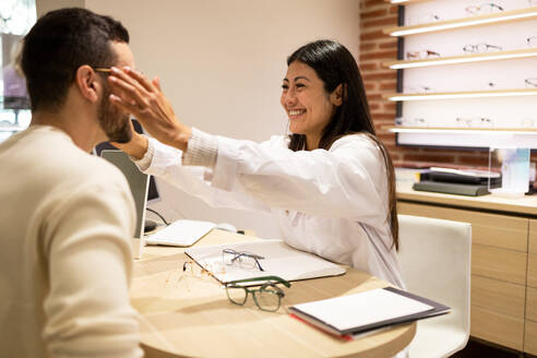Side view of young man smiling and applying optical eyeglasses with help of ophthalmologist while choosing eyewear in shop - ADSF44730