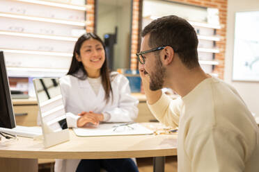 Side view of young male customer looking at mirror while selecting optical eyeglasses during consultation with eye doctor in store - ADSF44731