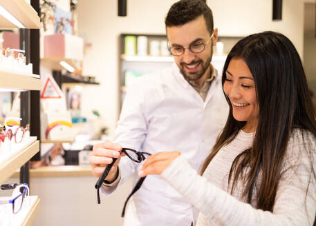 Smiling male specialist in white uniform helping female customer in buying new eyeglasses in optical store - ADSF44738
