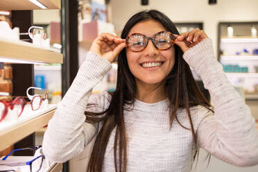 Positive female customer touching eyeglasses and looking at camera while selecting optical eyewear in store - ADSF44740