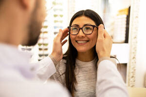 Side view of cropped anonymous male optician in uniform assisting smiling woman in trying different eyewear frames in store - ADSF44741