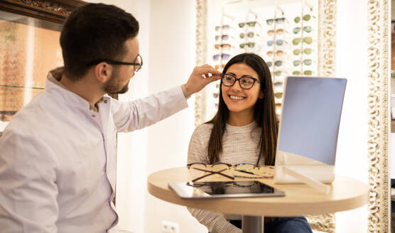 Male optician assisting smiling young female customer in trying different eyeglasses at table in optical shop - ADSF44742