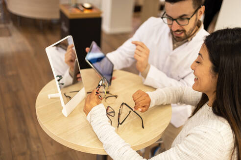 From above of smiling young woman in casual clothes choosing eyewear with the help of male optician holding digital tablet at store - ADSF44743