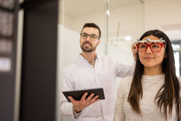 Male optician assisting young woman in casual clothes with long dark hair checking vision with trial frame in optical store - ADSF44744