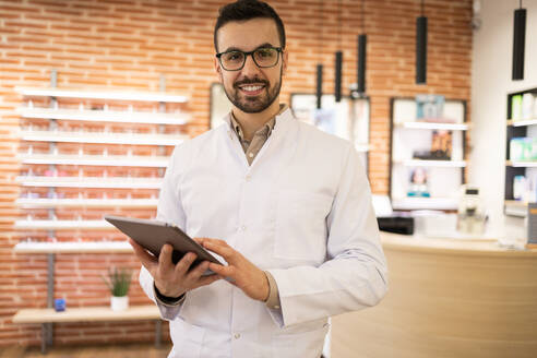 Portrait of smiling young male optician in white uniform and eyeglasses using digital table while looking at camera in optical shop - ADSF44745