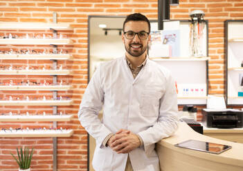 Positive young male optician in white uniform and glasses smiling and looking at camera while at counter in optical shop - ADSF44746