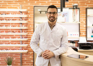 Positive young male optician in white uniform and glasses smiling and looking at camera while at counter in optical shop - ADSF44746