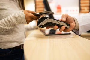 Side view of crop anonymous female customer paying contactless using mobile phone at counter at optical store - ADSF44747