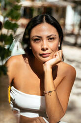 Confident young ethnic female in trendy outfit touching chin and looking at camera while resting in tropical resort - ADSF44748