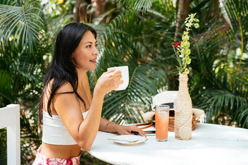 Full body of positive young mixed race female in summer outfit smiling and looking away while enjoying hot coffee in outdoor cafe of Tulum against tropical plants - ADSF44750