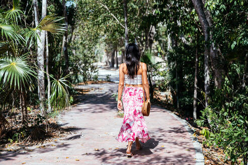 Full body of anonymous calm young mixed race female in colorful summer outfit with bare shoulders and bag walking on paved road among green exotic palms in Tulum - ADSF44754