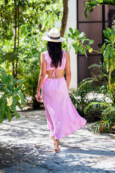 Calm anonymous young Mexican and Japanese female wearing pink summer maxi dress looking at camera while standing in sunny green forest with palms and trees in Tulum - ADSF44755