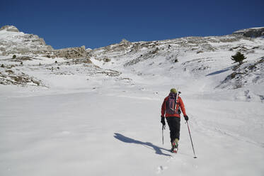 Back view of unrecognizable skier in warm ski suit and with backpack strolling along snowy mountain ridge against cloudless blue sky on ski resort in Pico Tortiellas, Canfranc, Spain - ADSF44784